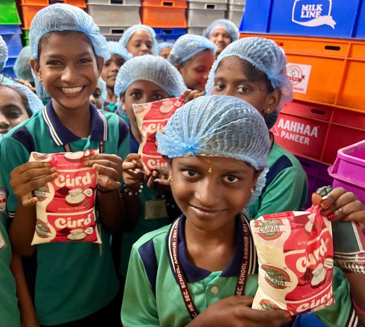 Students wearing hairnets holding curd packets during an Aavin dairy plant educational visit